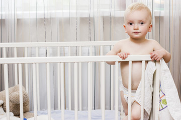 toddler boy standing in his crib holding a blanket serious