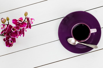 cup of tea or coffee on violet plate, silver tea spoon, orchid flower on white colored wooden table,  top view