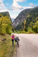 Fototapeta na wymiar Young woman with backpack and bicycle walking on countryside terrain. Tourist emerging from a lengthy uphill, mountains in the background