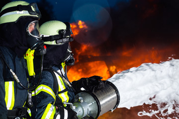 Naklejka premium Feuerwehr - Feuerwehrmänner löschen ein großes Feuer, sie stehen vor einer Feuerwand und tragen Schutzkleidung