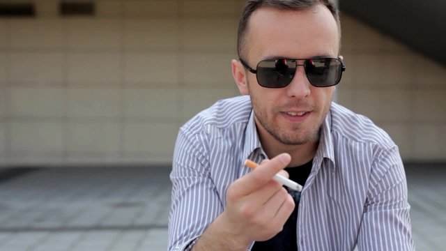 Young, handsome man in sunglasses and blue shirt, sitting somwhere on the street and smmoking cigarette. He is probably talking with someone