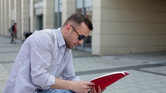 Handsome man  in blue shirt and sunglasses, sitting near building and reading notes. He is talking to someone.