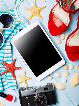 Summer women's accessories: sunglasses, red shoes, dress, tablet, camera, on blue wood background.