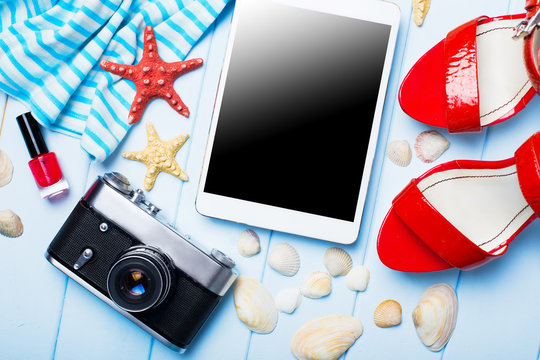 Summer women's accessories: sunglasses, red shoes, dress, tablet, camera, on blue wood background.
