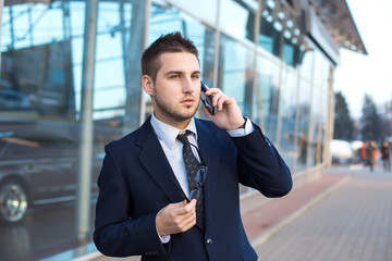 Attractive young businessman speak a cell phone, smiling. Standing  in front of a modern office building.