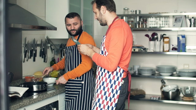 Two male friends cooking and taking photo with cellphone in kitchen
