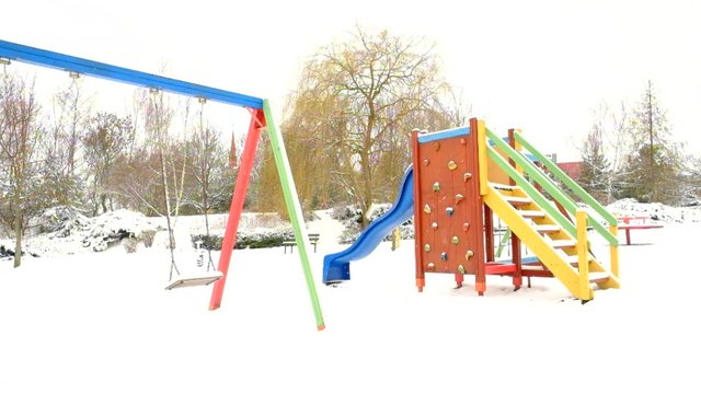 Children playground with seesaw, slider, wooden ladder, turning merry-go-round. Abandoned playground in winter under snow at park, slowly snowing. 