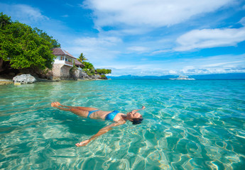 young woman in bikini relaxing lying on water against background beach and bungalow