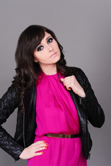 Fototapeta premium Stylish girl 20-25 year old wearing trendy leather jacket and pink dress posing in studio over gray. Looking at camera. Young adults. Spring season. 20s.
