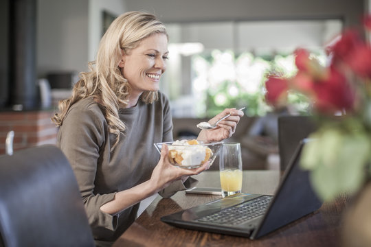 Woman eating fruit salad working at home on laptop