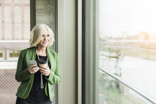 Portrait of blond businesswoman with coffee to go and smartphone looking through window
