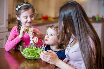 Happy family eating fresh green salad together around wooden table at home. Mother and her daughters enjoying family healthy lunch together. Healthy lifestyle.