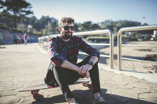 Spain, La Coruna, portrait of hipster wearing sunglasses sitting on his longboard