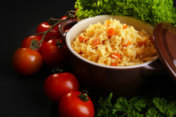 Stewed rice with a carrot and tomatoes in a cooking pot over black background, close up