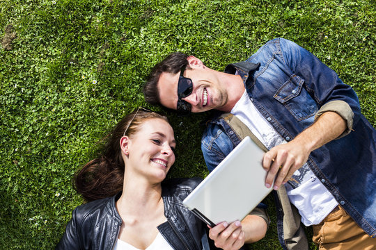 Happy young couple lying in meadow looking at digital tablet