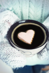Female hands in warm mittens holding cup of hot coffee with heart marshmallow, close up