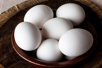 White eggs in a brown clay cracked bowl on wooden old weathered cracked cutting board on rough beige rural map closeup from above