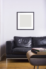 A black picture frame hanging on a white wall.  Interior scene also has leather sofa, wooden flooring and a light wood table.