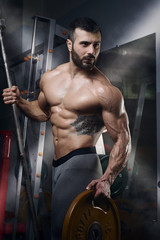 Men's physique athlete standing with the barbell and  the weight plate in the gym