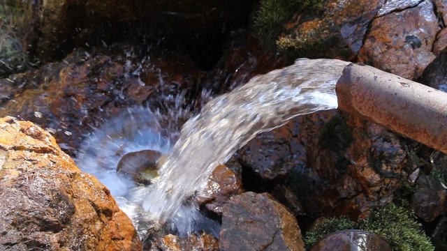 Transparent and pure cold water flows from a metal pipe and breaks about stones
