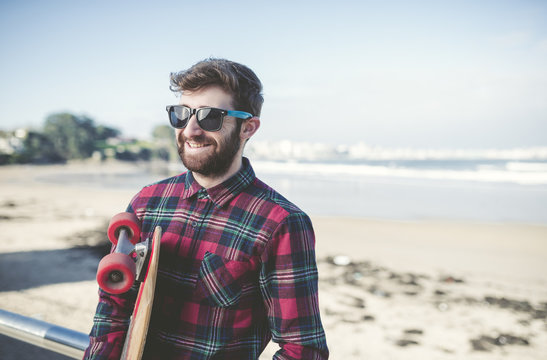 Spain, La Coruna, portrait of smiling hipster wearing sunglasses with his skateboard on the beach