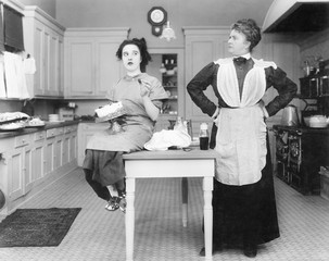 Housekeeper in the kitchen glaring at a young woman eating a cake 