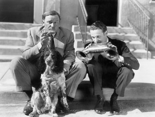 Two men playing two harmonicas with an English Setter 