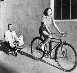 Woman on a bicycle pulling a grown man on a toy tricycle 