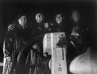 Four men in slickers standing together at the captain's wheel of a ship 