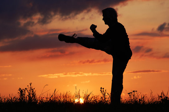 Man practicing karate on the grassy horizon after sunset. Karate kick leg. Art of self-defense. Silhouette on a background of dramatic clouds at sunset.