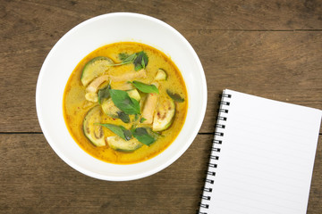The delicious homemade Thai green curry with pork (Gang Keaw Wan) in white bowl and blank note book on old wooden planks.