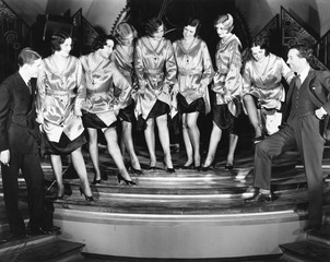 A chorus line of women showing off their legs to two men 