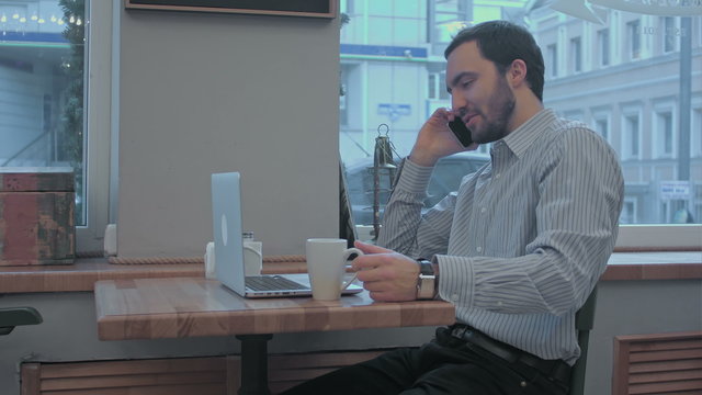 Handsome businessman speaking on phone and working with laptop in cafe