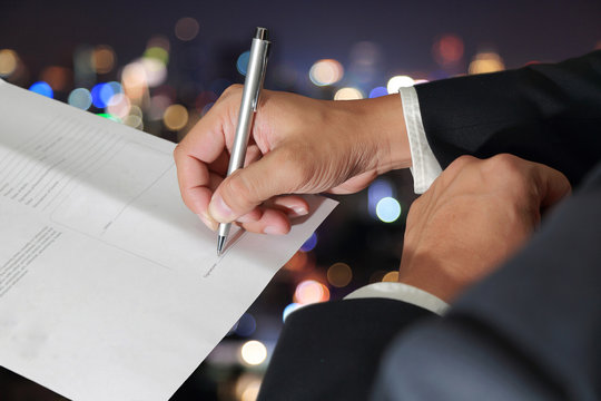 Businessman's Hand holding a pen signing to the document on blurred abstract night bokeh background as Approval Concept.