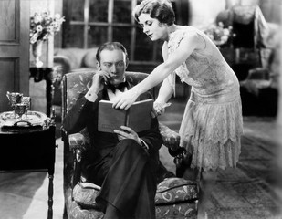 Profile of a young woman showing a book to a man sitting in an armchair 