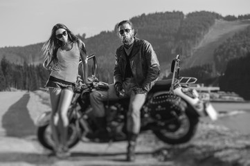 Fototapeta na wymiar Couple standing by a motorbike. Handsome man wearing leather jacket, gloves and boots and young beautiful sexy woman wearing shorts. Summer day. Tilt lens blur effect. Black and white