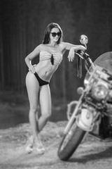Obraz na płótnie Canvas Sexy girl with long hair wearing swimming suit and high heels standing by the motorcycle and smiling. Motorcycle is standing on the side of the road. Tilt shift lens blur effect. Black and white