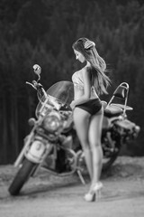 Fototapeta na wymiar Beautiful sexy girl with long hair wearing swimming suit and high heels standing by the motorcycle and looking down on the side of the road in the mountains. Tilt lens blur effect. Black and white