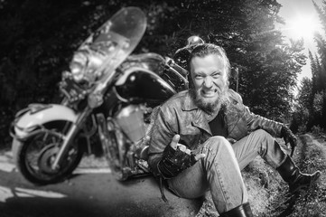 Unshaven male biker in leather jacket and jeans sitting on road near motorcycle and giving the...