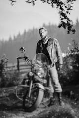 Young handsome biker standing by his custom made cruiser motorcycle on a sunny day with forest on the background. Tilt shift lens blur effect. Black and white