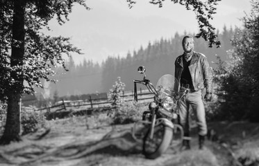 Young handsome biker standing by his custom made cruiser motorcycle on a sunny day with forest on the background and smiling. Horizontal picture. Tilt shift lens blur effect. Black and white
