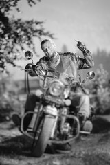 Naklejka premium Handsome biker with beard driving his cruiser motorcycle in the forest and giving the devil horns gesture and smilling. Man is wearing leather jacket and jeans. Tilt lens blur effect. Black and white