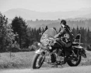 Fototapeta na wymiar Long-haired biker in sunglasses and a leather jacket sitting on a black custom motorcycle and looking into distance. Sunny day in the mountains. View from the back. Tilt blur effect. Black and white