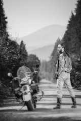 Obraz premium Handsome biker with long hair wearing leather jacket, jeans, boots and gloves standing near his custom made cruiser motorcycle on the open road. Looking to the sun. Tilt soft effect. Black and white