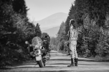 Obraz premium Biker with long hair wearing leather jacket jeans leather boots and gloves standing near his custom made cruiser motorcycle on the open road. Looking into distance. Tilt soft effect. Black and white