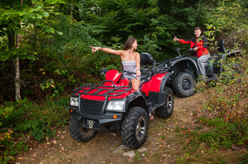Fototapeta na wymiar Happy beautiful couple driving four-wheelers ATV and looking at each other. Girl is pointing into the distance. Man is giving thumb up
