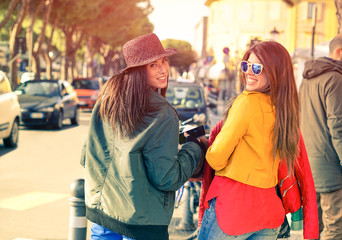 Beautiful women looking back to camera while walking in old city center - Best friends girls with casual style holding phone and red jacket - Vintage filter look with sun halo -Focus on right  girl - obrazy, fototapety, plakaty