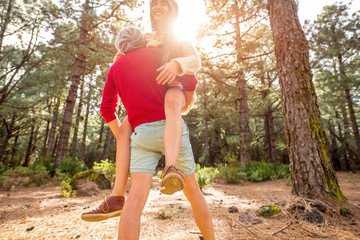 Young playful couple in sweaters and hats having a piggyback ride in the pine forest. Strong...