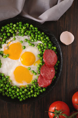 Fried eggs with sausage and green peas on a black frying pan on a wooden background