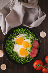 Fried eggs with sausage and green peas on a black frying pan on a wooden background
